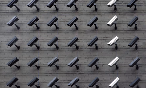 A security camera mounted on a residential wall with a digital scanning overlay on a human face, representing intrusive biometric surveillance.